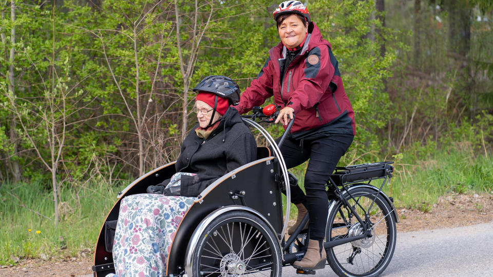 Kvinna som jobbar inom äldreomsorgen skjutsar äldre kvinna på en parcykel.