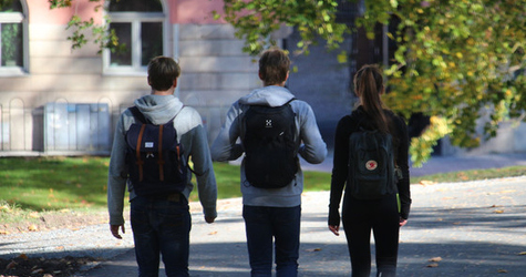 Tre ungdomar med ryggsäckar promenerar  längs en väg en somrig dag.